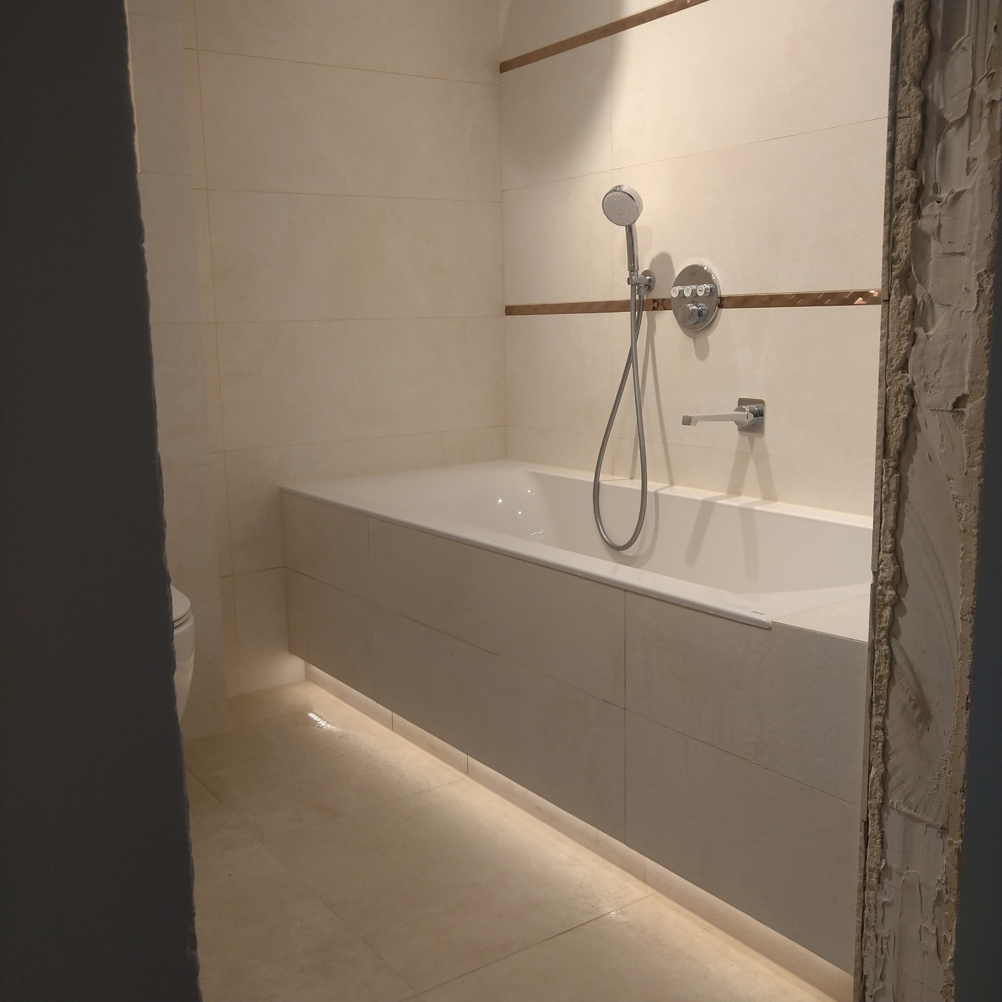 Nowoczesna łazienka z wanną obudowaną płytkami, armaturą i subtelnym oświetleniem pod wanną, widok z perspektywy wejścia z fragmentem surowej ściany.