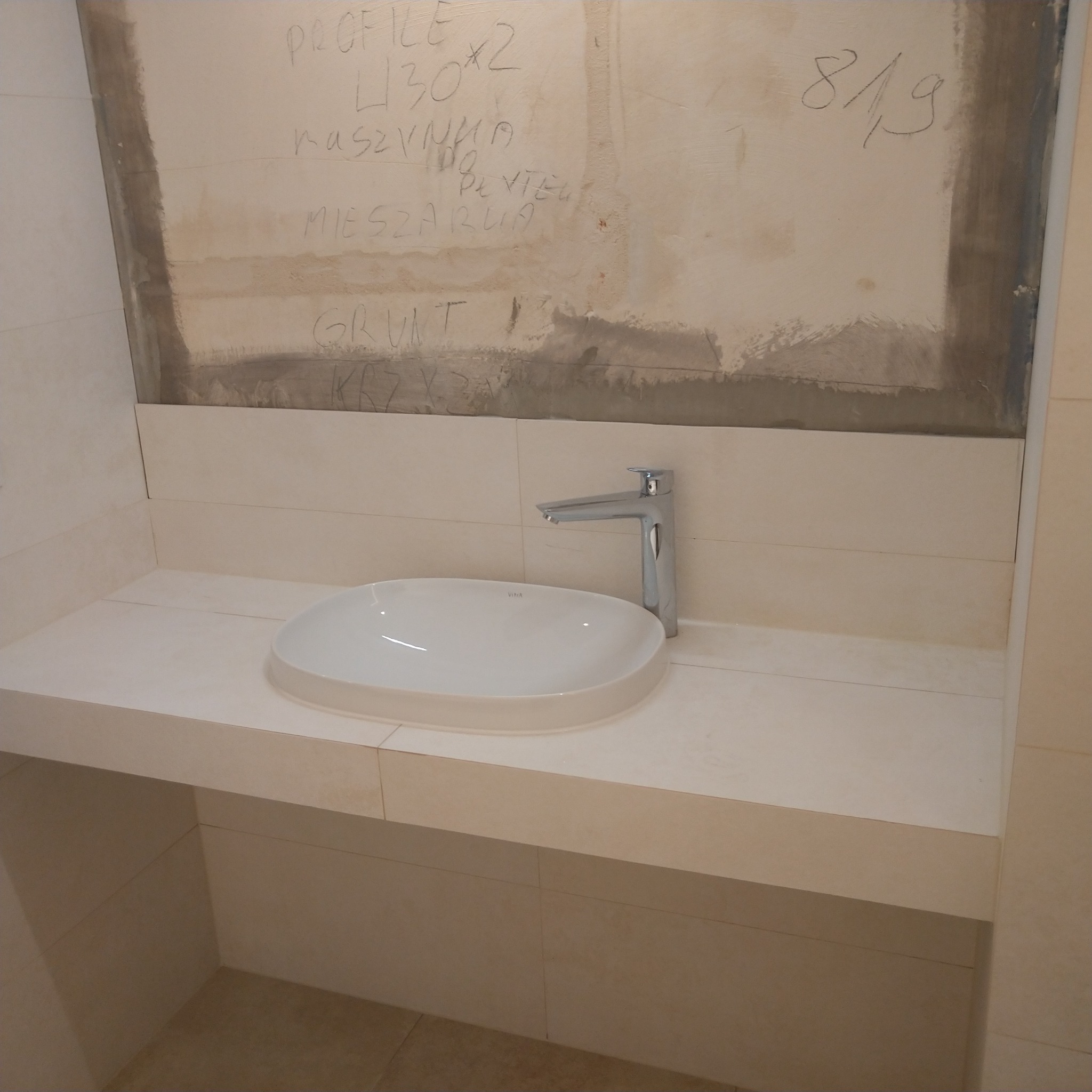 Umywalka nablatowa z chromowaną baterią, zamontowana na blacie z płytek w łazience w trakcie remontu, widoczne notatki na ścianie nad płytkami.