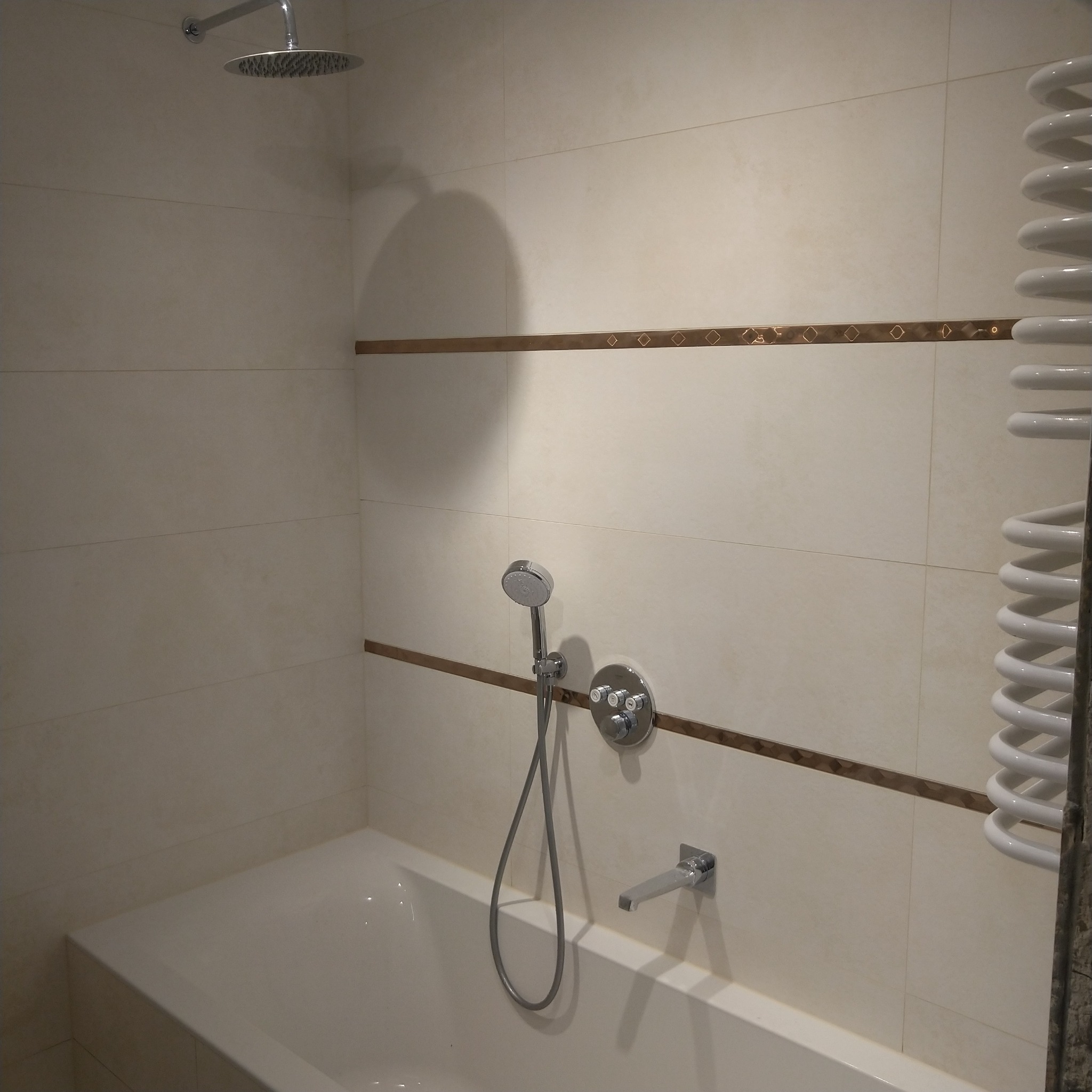 Jasna łazienka z beżowymi płytkami, dekoracyjnym bordiurem, baterią wannową, słuchawką prysznicową i grzejnikiem drabinkowym.