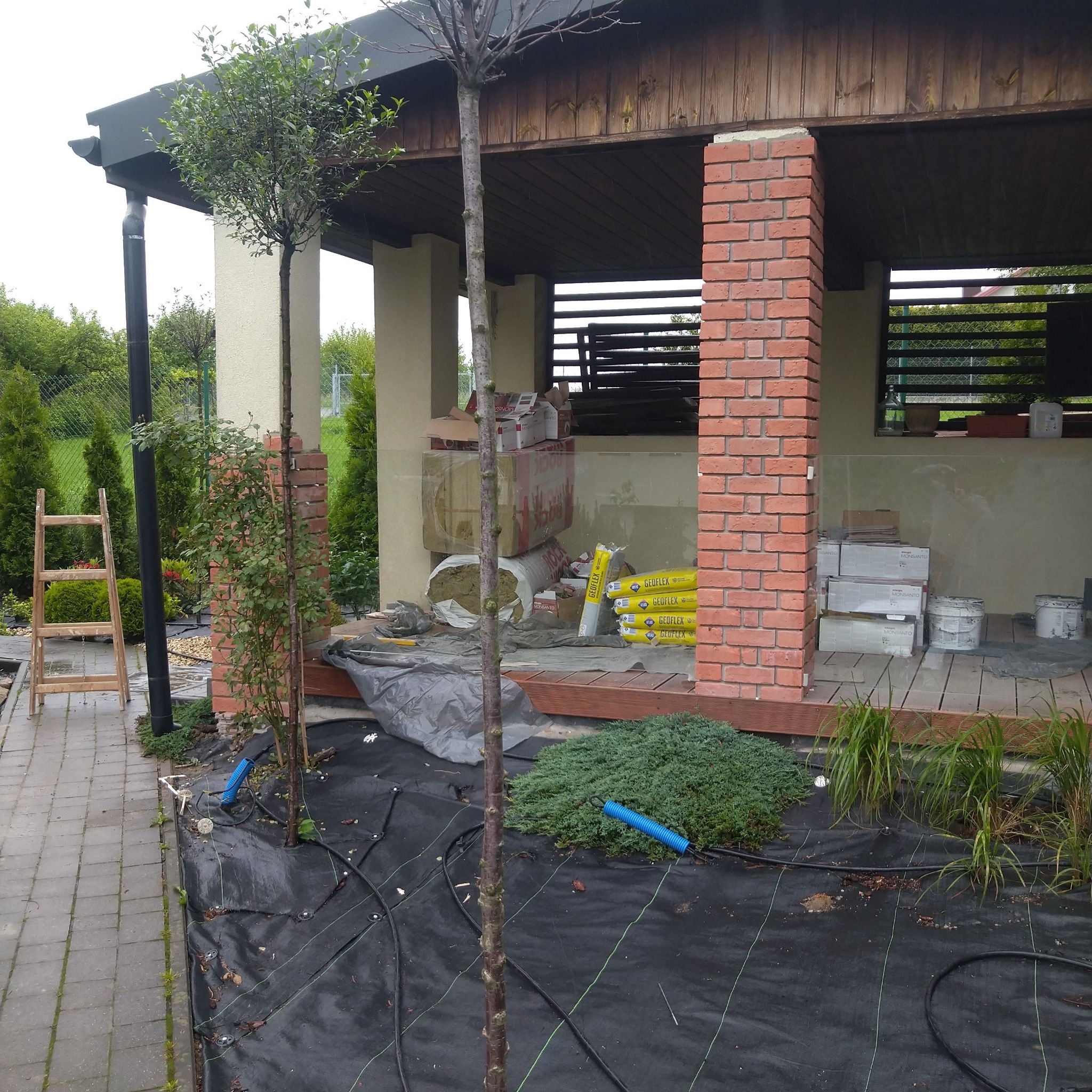 Przygotowania do ocieplenia poddasza w altanie ogrodowej: worki z wełną mineralną Geoflex, płyty Monsanto, ceglane filary, drewniana podłoga i czarna agrowłóknina z systemem nawadniania...