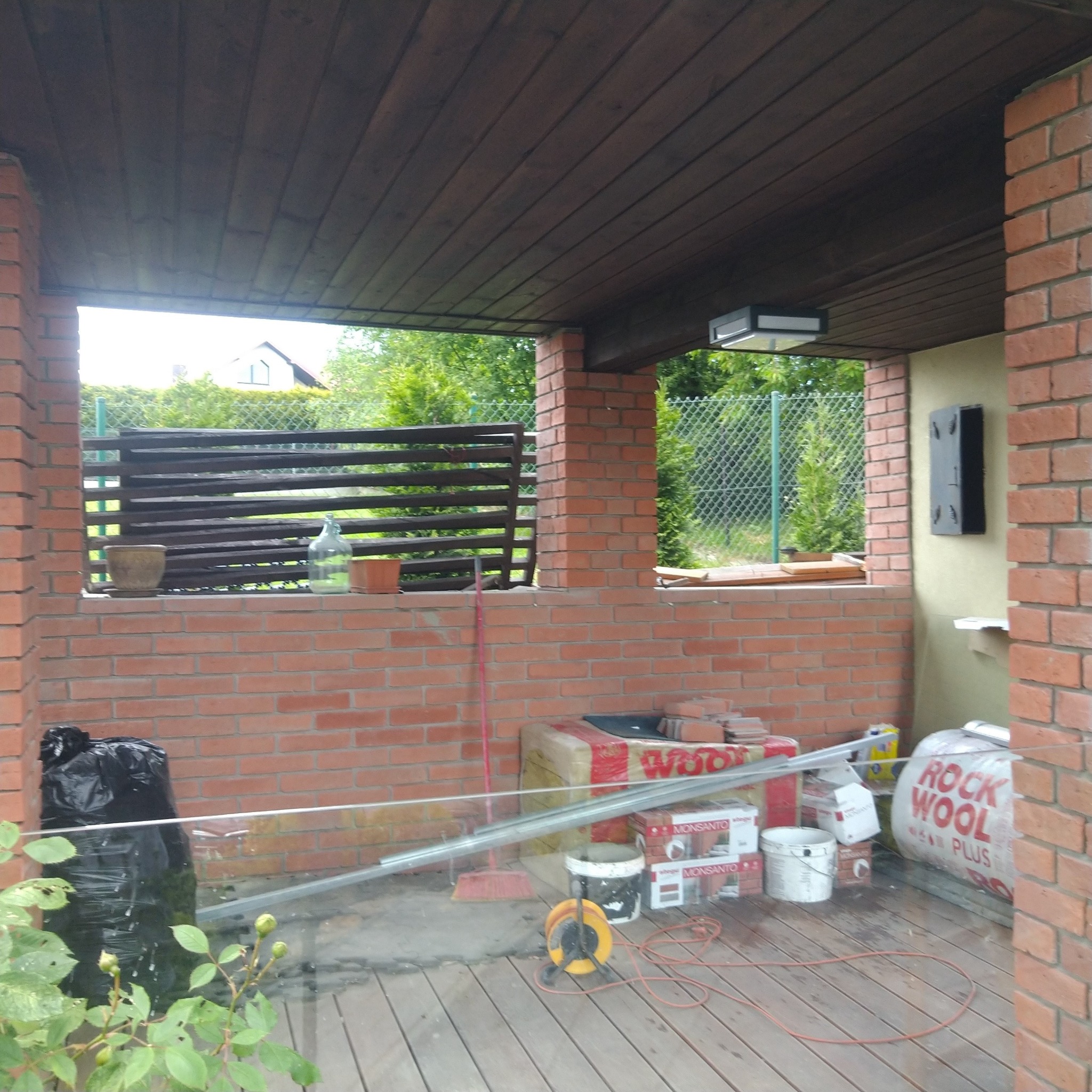 Przygotowania do prac budowlanych na zadaszonym tarasie: paczki wełny mineralnej Rockwool, puszki farb, przedłużacz, profil aluminiowy, ceglana konstrukcja z drewnianym zadaszeniem, widok na ogród.