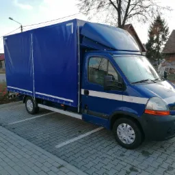 Niebieski samochód dostawczy z plandeką zaparkowany na parkingu z kostki brukowej; widoczny numer '7' na plandece i zabudowa nad kabiną kierowcy.