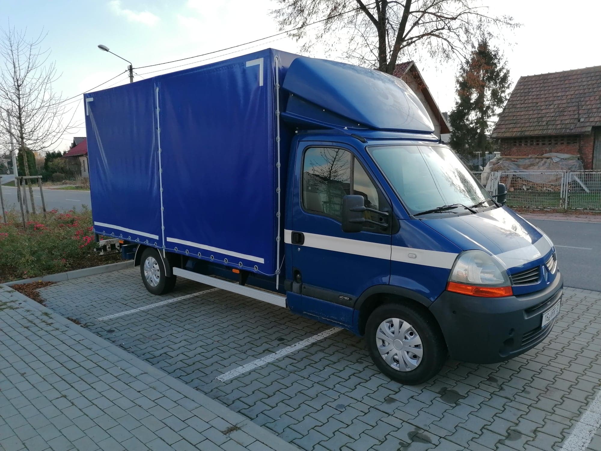 Niebieski samochód dostawczy z plandeką zaparkowany na parkingu z kostki brukowej; widoczny numer '7' na plandece i zabudowa nad kabiną kierowcy.