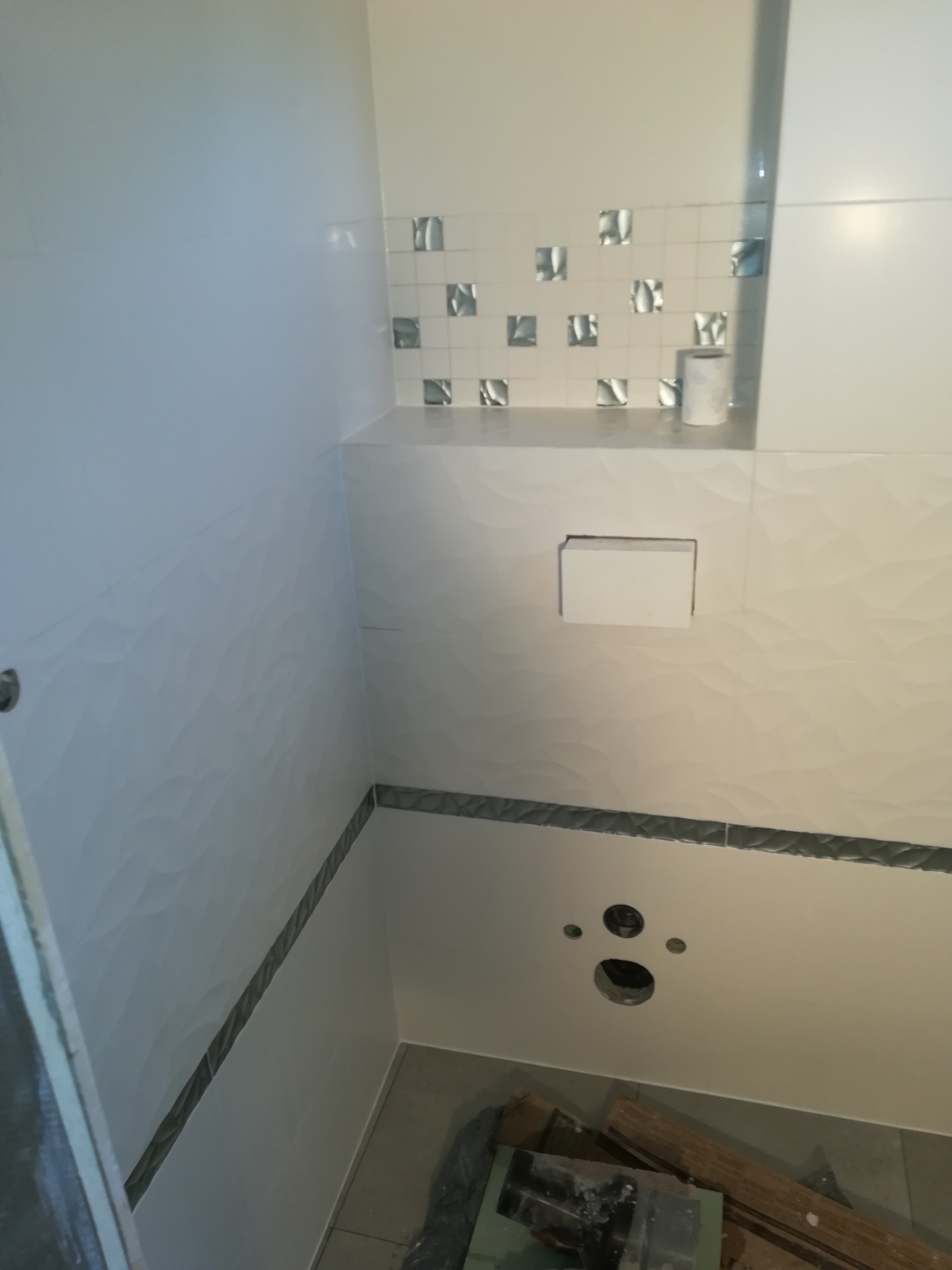 Ściana w łazience wyłożona białymi płytkami z delikatnym wzorem fal, u góry mozaika z kwadratowych płytek z metalicznym wzorem, widoczne otwory na armaturę i fragment podłogi z pozostałościami...