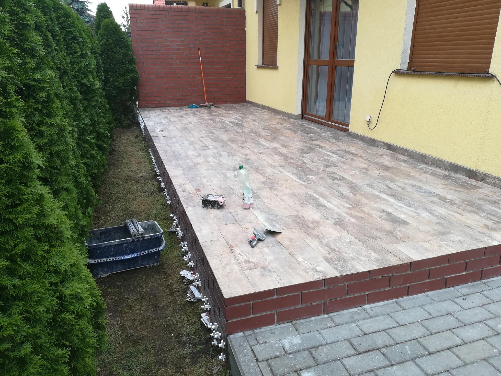 Wykończony taras z płytek imitujących drewno, obłożony cegłą klinkierową, widoczne krzyżyki dystansowe, wiadro z zaprawą i narzędzia na powierzchni, zieleń w tle.