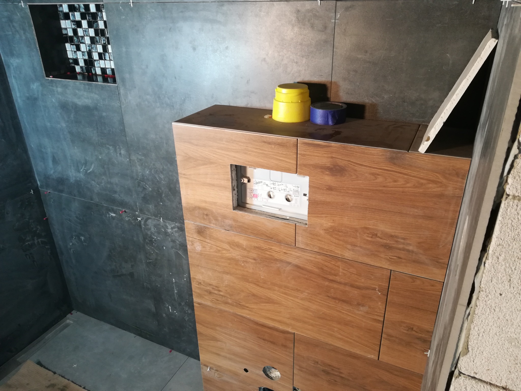 Montaż płytek w łazience: ciemnoszare płytki na ścianach z poziomymi fugami, mozaika czarno-biała w niszy, obudowa stelaża WC z płytek imitujących drewno, żółty pojemnik i niebieska taśma na obudowie.