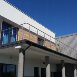 Nowoczesny dom z balkonem zabezpieczonym stalową balustradą, widok z dołu na tle błękitnego nieba.