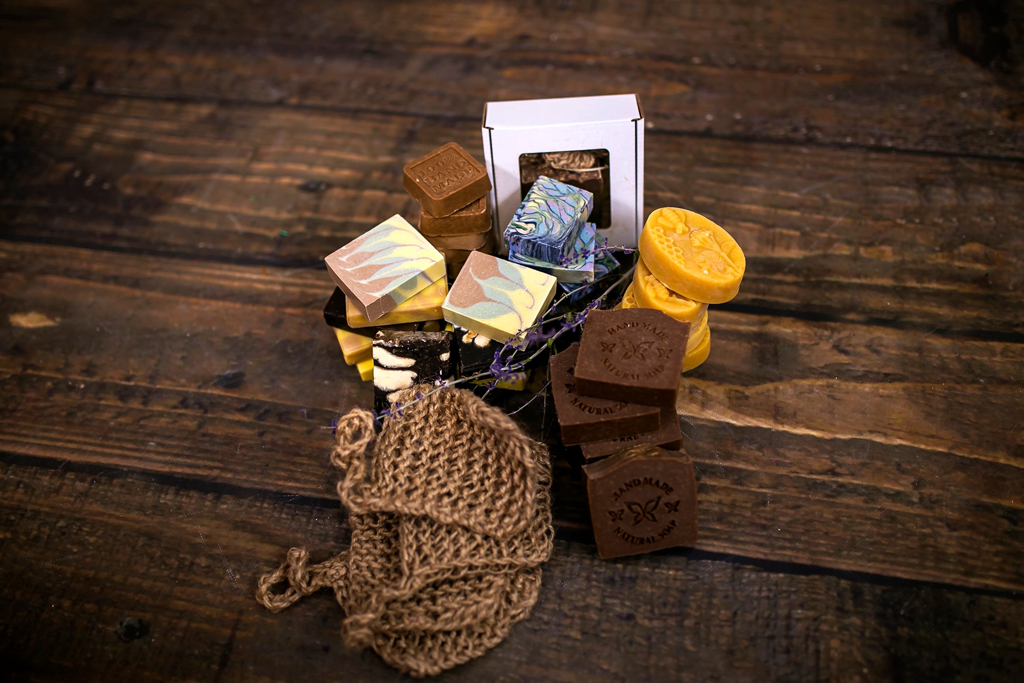 Różnorodne, ręcznie robione mydła w kostkach i w formie plastrów miodu, ułożone na drewnianym stole obok jutowego woreczka do mycia, prezentowane w naturalnym świetle.