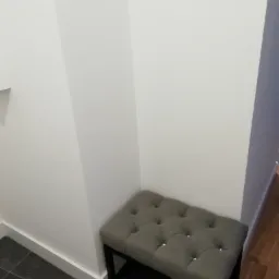 Szary puf z pikowaniem w rogu pokoju, na ciemnej podłodze, przy białej ścianie z listwą przypodłogową.