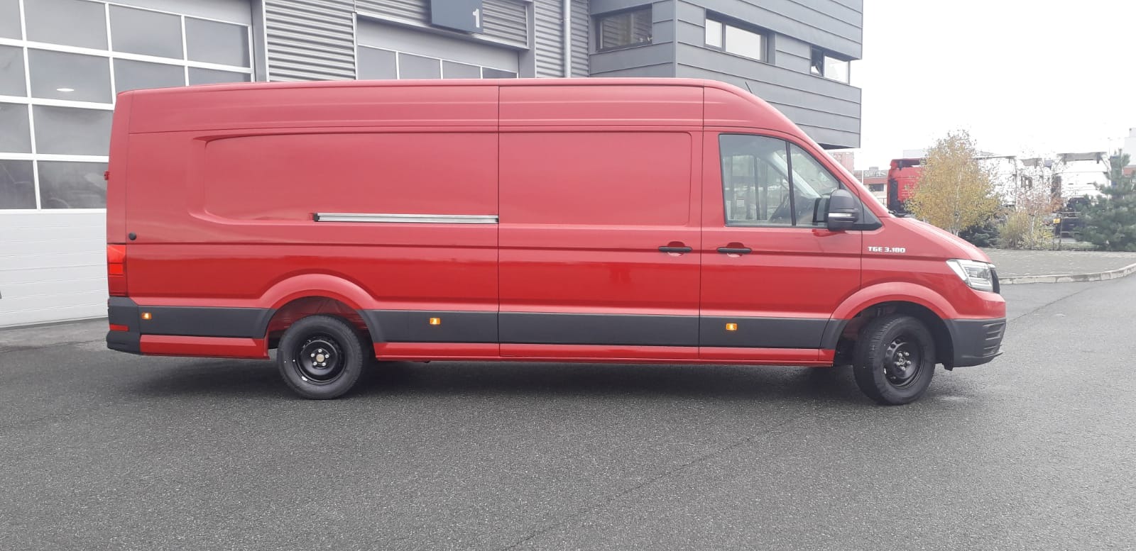 Czerwony, dostawczy van marki MAN TGE 3.180 z wysokim dachem, zaparkowany przed nowoczesnym budynkiem z szarą elewacją i dużymi oknami.