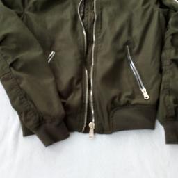 Renata Niemiec - Oliwkowa kurtka bomberka z metalowymi zamkami błyskawicznymi, leżąca na białym tle.