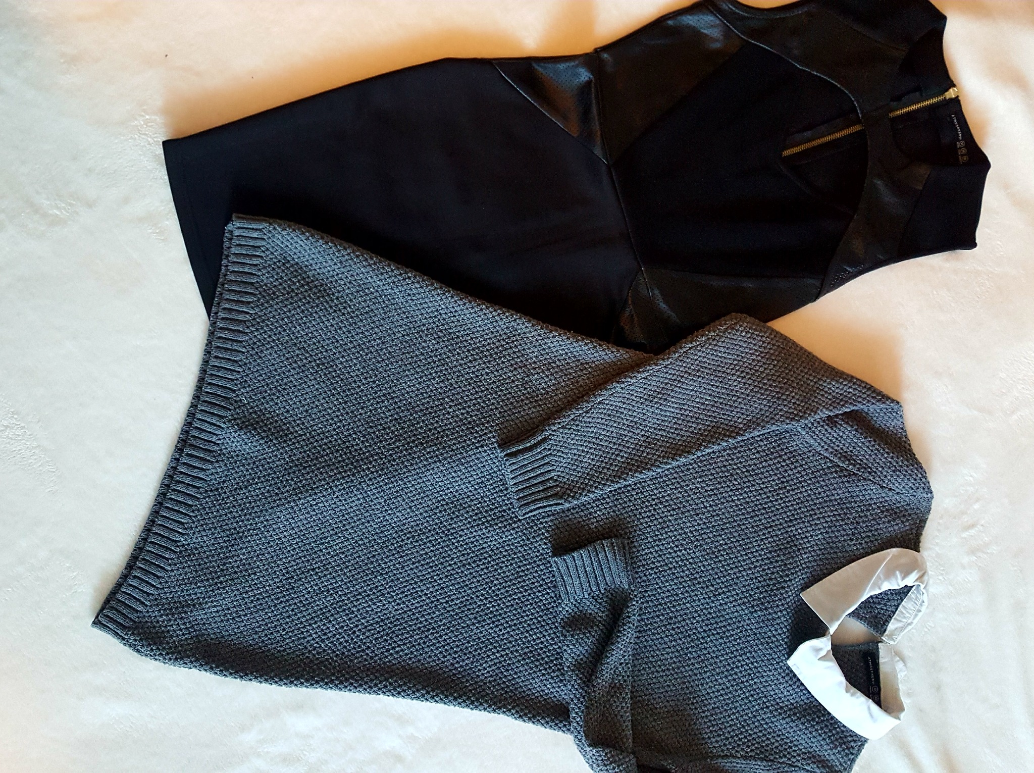 Dwa ubrania damskie leżące na jasnym tle: czarna sukienka z elementami imitującymi skórę i złotym zamkiem z tyłu oraz szary sweter z białym kołnierzykiem.