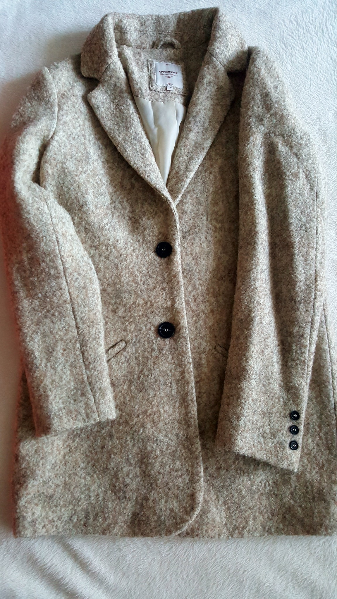 Beżowy, wełniany płaszcz damski z dwoma czarnymi guzikami, widoczna podszewka i metka, leżący na białym, puszystym tle.