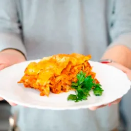 Lasagne na białym talerzu, udekorowana świeżą zieloną pietruszką, prezentowana przez osobę w szarym swetrze na tle jasnego wnętrza.
