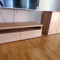 Drewniana szafka RTV z dwoma szufladami i półką na sprzęt, obok szafa z jasnego drewna, parkiet na podłodze, telewizor na ścianie.