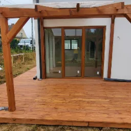 Nowo wybudowany taras z drewna z drewnianą pergolą, widoczny pracownik w kraciastej koszuli, biała ściana budynku i materiały budowlane obok.