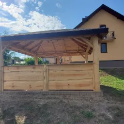 Nowoczesna, drewniana konstrukcja z naturalnych bali, częściowo osłonięta deskami, na tle domu jednorodzinnego i błękitnego nieba z chmurami.