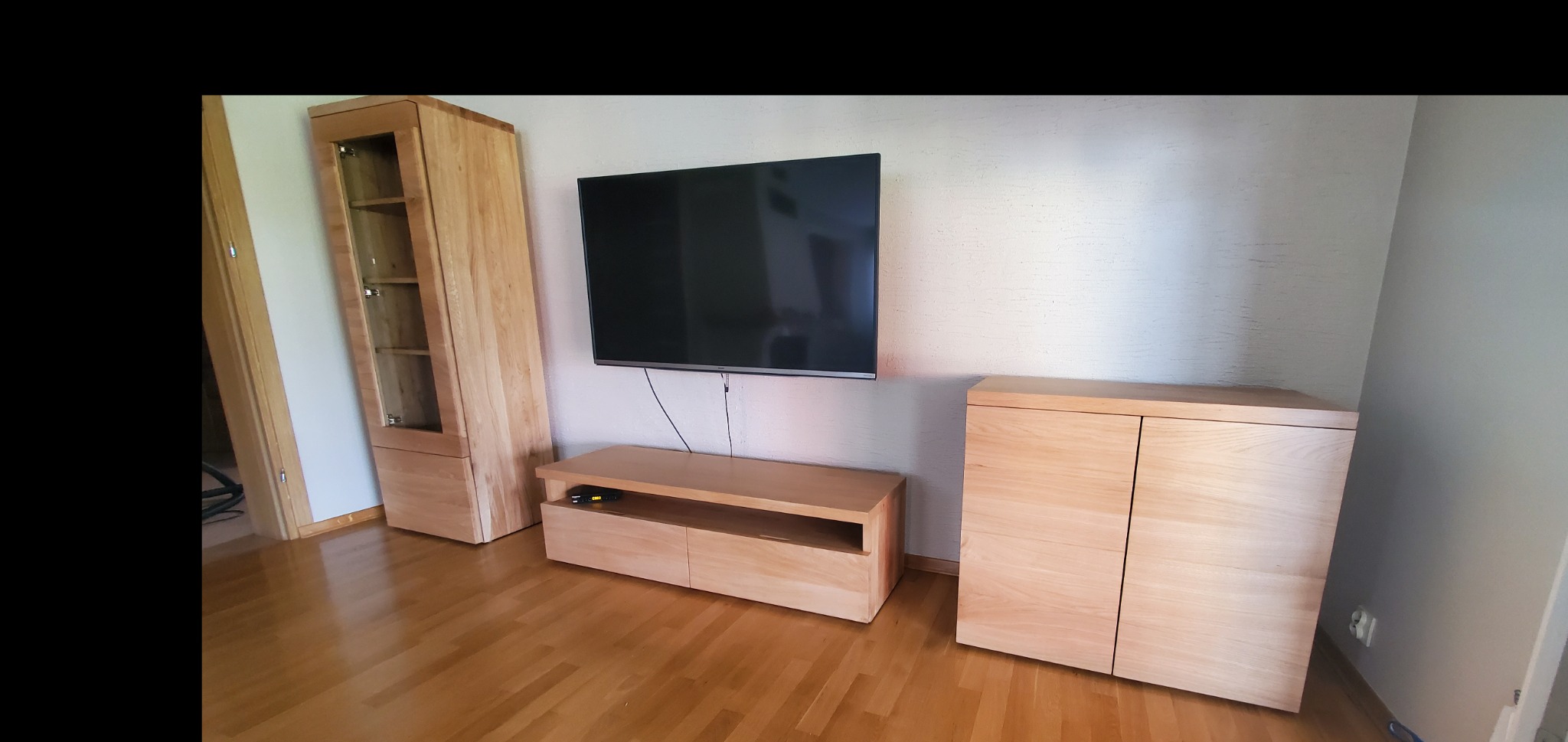 Jasne drewniane meble do salonu: witryna z przeszklonymi drzwiami, szafka RTV i komoda z dwojgiem drzwi, ustawione na parkiecie pod wiszącym telewizorem.