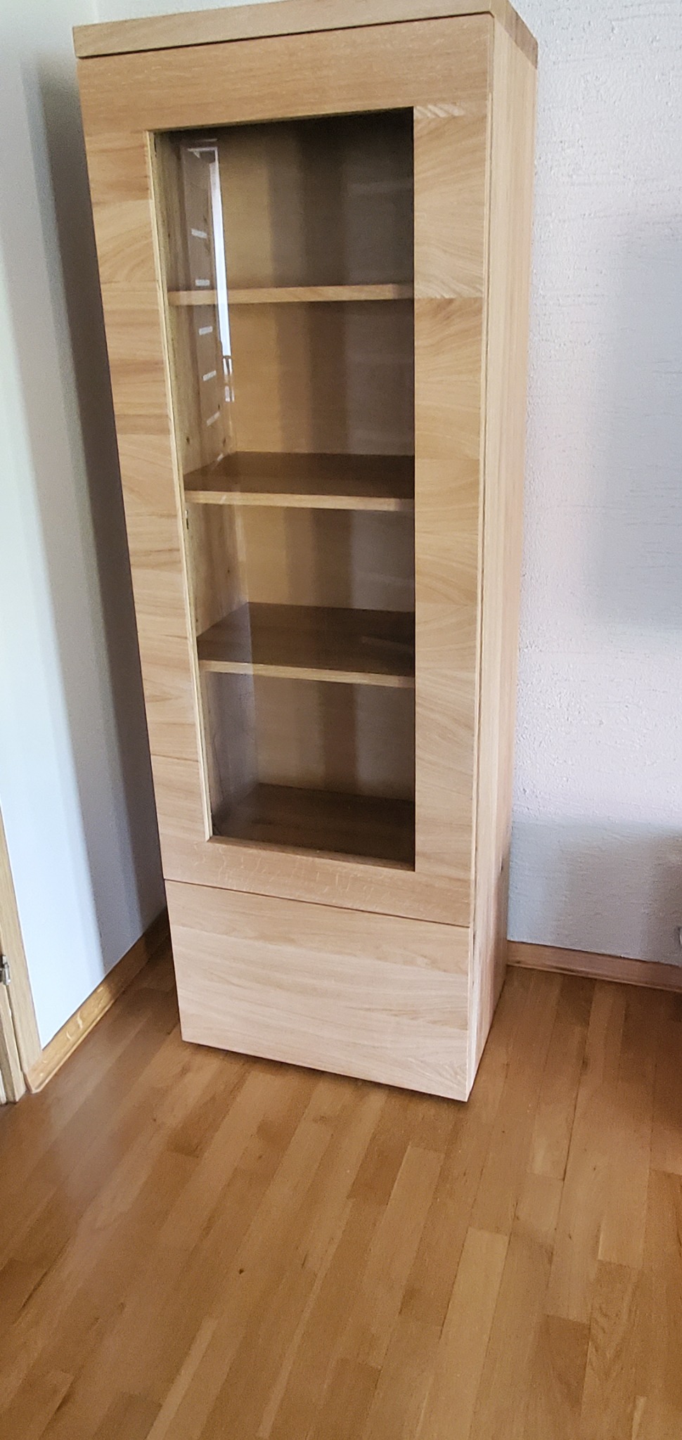 Jasna, drewniana witryna z przeszklonymi drzwiami i półkami, stojąca na parkiecie w minimalistycznym wnętrzu.