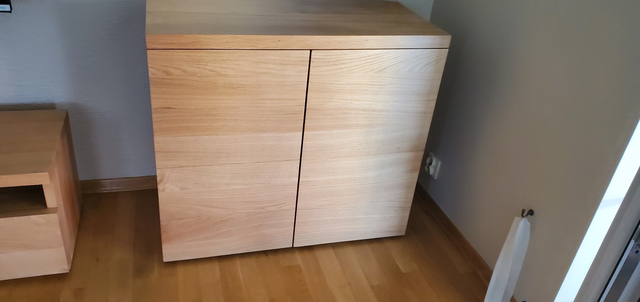 Dwudrzwiowa szafka z jasnego drewna o minimalistycznym designie, z widoczną strukturą drewna, stojąca na drewnianej podłodze w jasnym wnętrzu.