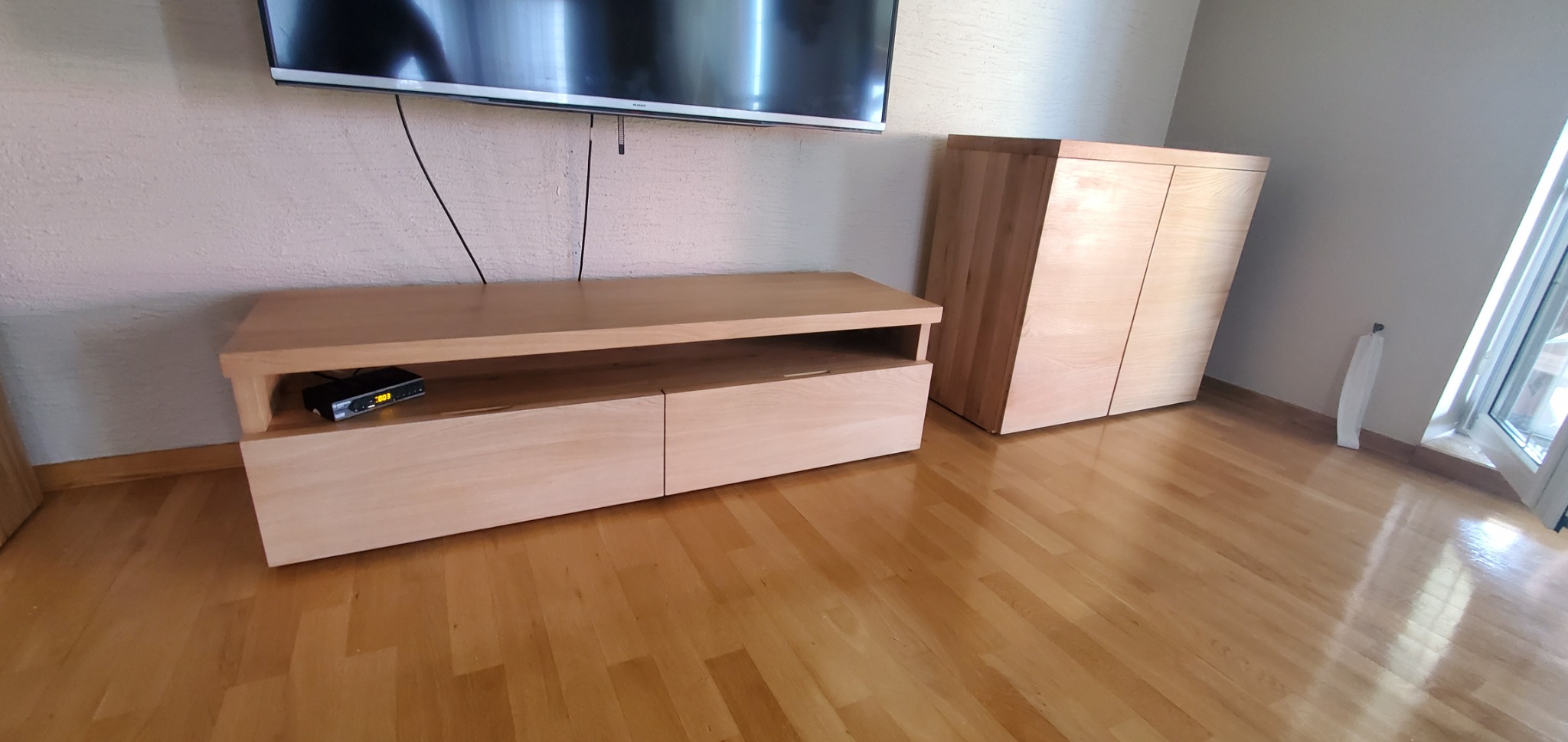 Drewniana szafka RTV z dwoma szufladami i półką na sprzęt, obok szafa z jasnego drewna, parkiet na podłodze, telewizor na ścianie.