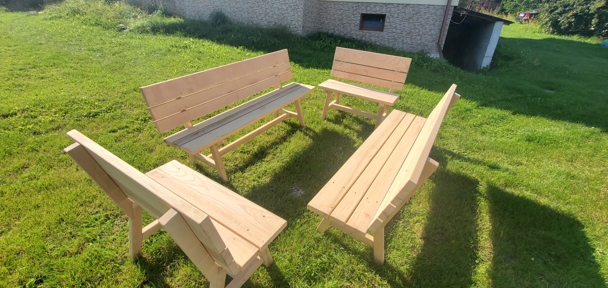 Zestaw surowych, drewnianych ławek z oparciami, ustawionych w okręgu na trawniku przed domem z cegły.