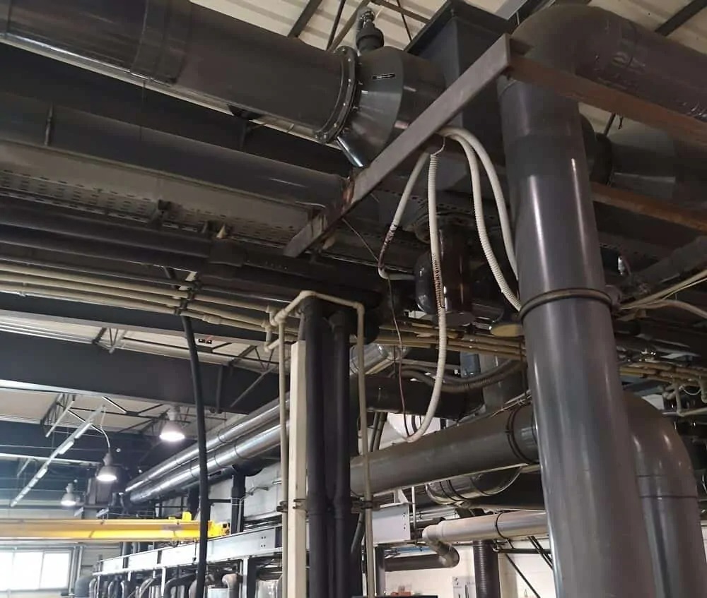 Szare rury wentylacyjne i instalacje w hali przemysłowej, widoczne kable i oświetlenie sufitowe.