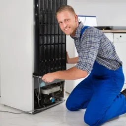 Uśmiechnięty mężczyzna w niebieskim kombinezonie, klęczący obok białej lodówki, używa klucza do dokręcenia elementu w dolnej części urządzenia.