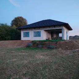 Surowy stan parterowego domu z ciemnym dachem na wzniesieniu, okna zabezpieczone folią, widoczne ślady opon i hałda ziemi w tle, wczesny wieczór.