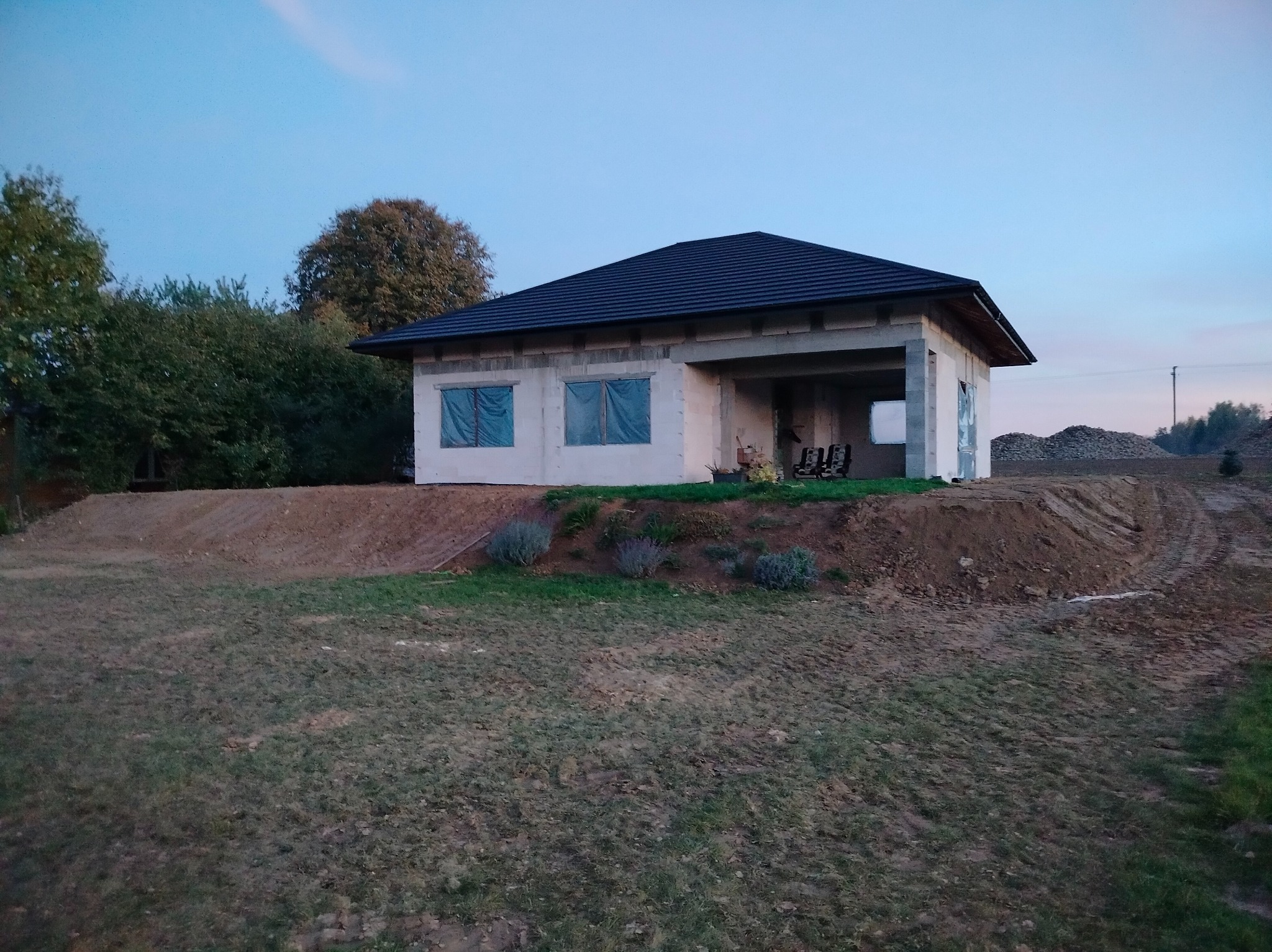 Surowy stan parterowego domu z ciemnym dachem na wzniesieniu, okna zabezpieczone folią, widoczne ślady opon i hałda ziemi w tle, wczesny wieczór.