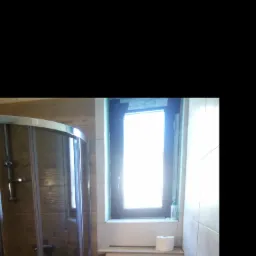 Wykończona łazienka z kabiną prysznicową z przesuwnymi drzwiami, brązowymi płytkami imitującymi drewno, oknem z czarną ramą i toaletą w zabudowie z chromowanym przyciskiem spłukiwania oraz rolką...