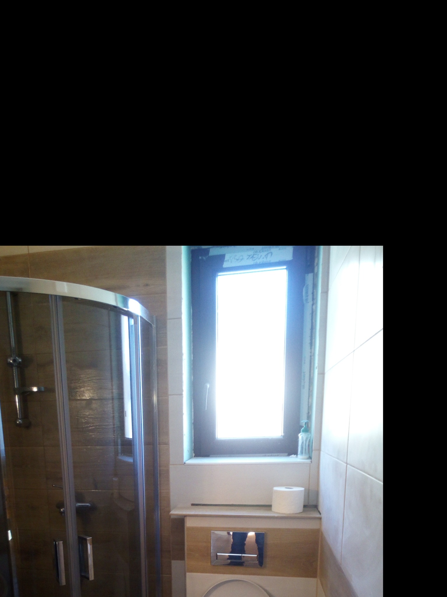 Wykończona łazienka z kabiną prysznicową z przesuwnymi drzwiami, brązowymi płytkami imitującymi drewno, oknem z czarną ramą i toaletą w zabudowie z chromowanym przyciskiem spłukiwania oraz rolką...