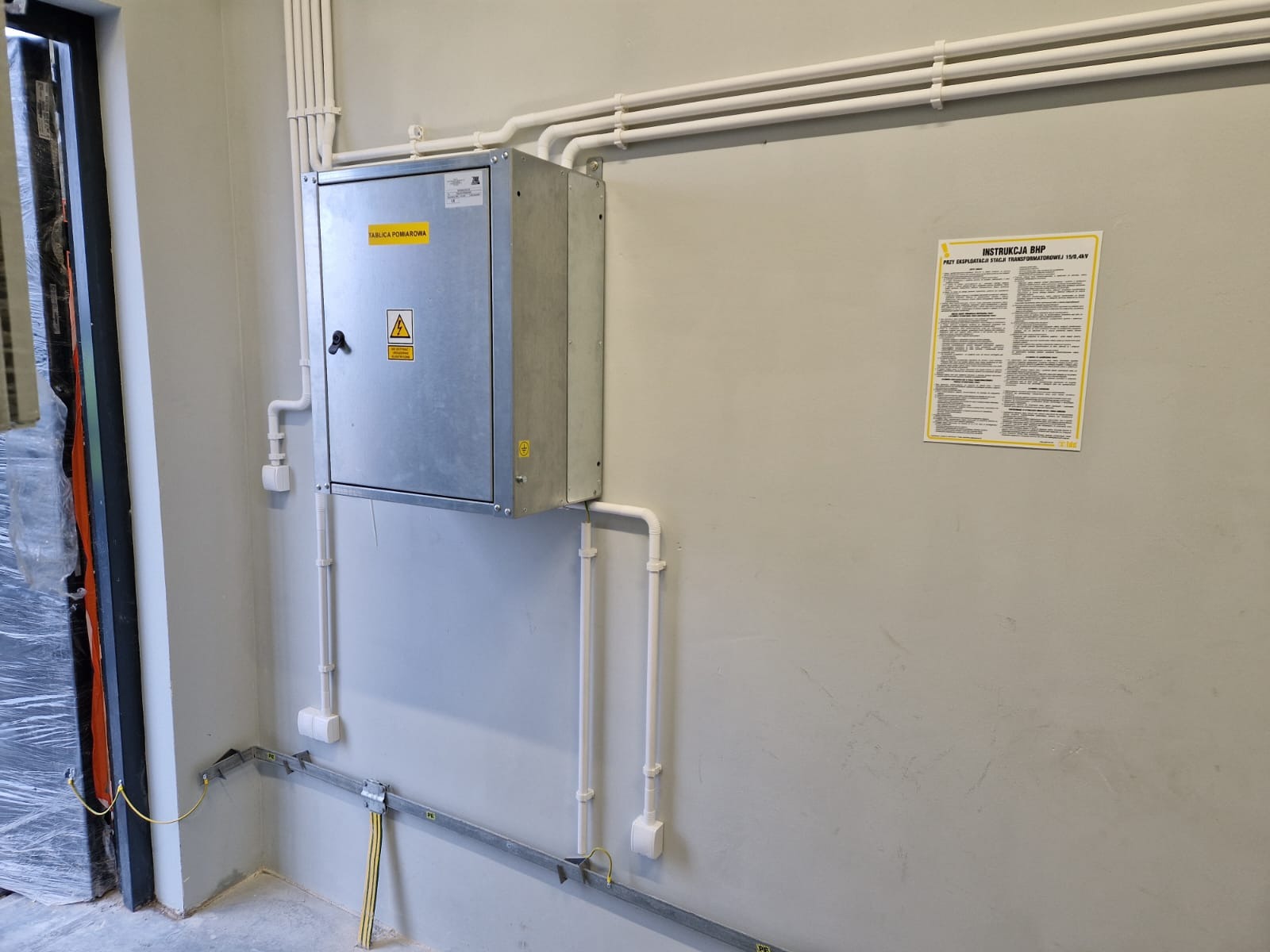 Metalowa skrzynka pomiarowa z naklejką ostrzegawczą i Instrukcją BHP na szarej ścianie, widoczne rury i kable w białej osłonie.