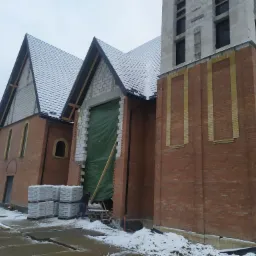 Budynek w trakcie budowy z cegły i bloczków, ocieplony fragment elewacji zabezpieczony zieloną folią, palety z materiałami budowlanymi na zewnątrz, zimowy krajobraz.