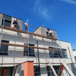 Ocieplanie i tynkowanie elewacji budynku wielorodzinnego z użyciem rusztowań, widoczni pracownicy podczas pracy, błękitne niebo w tle.