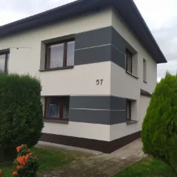 Dwukondygnacyjny dom po renowacji elewacji, z numerem '57', pomalowany w odcieniach bieli i szarości, z brązowymi oknami i ciemną podmurówką, otoczony zielenią.