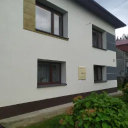 Dwupiętrowy dom z jasną elewacją, brązowymi oknami i szarą dekoracją, skrzynka gazowa i numer 57 na ścianie, widok z poziomu ogrodu.