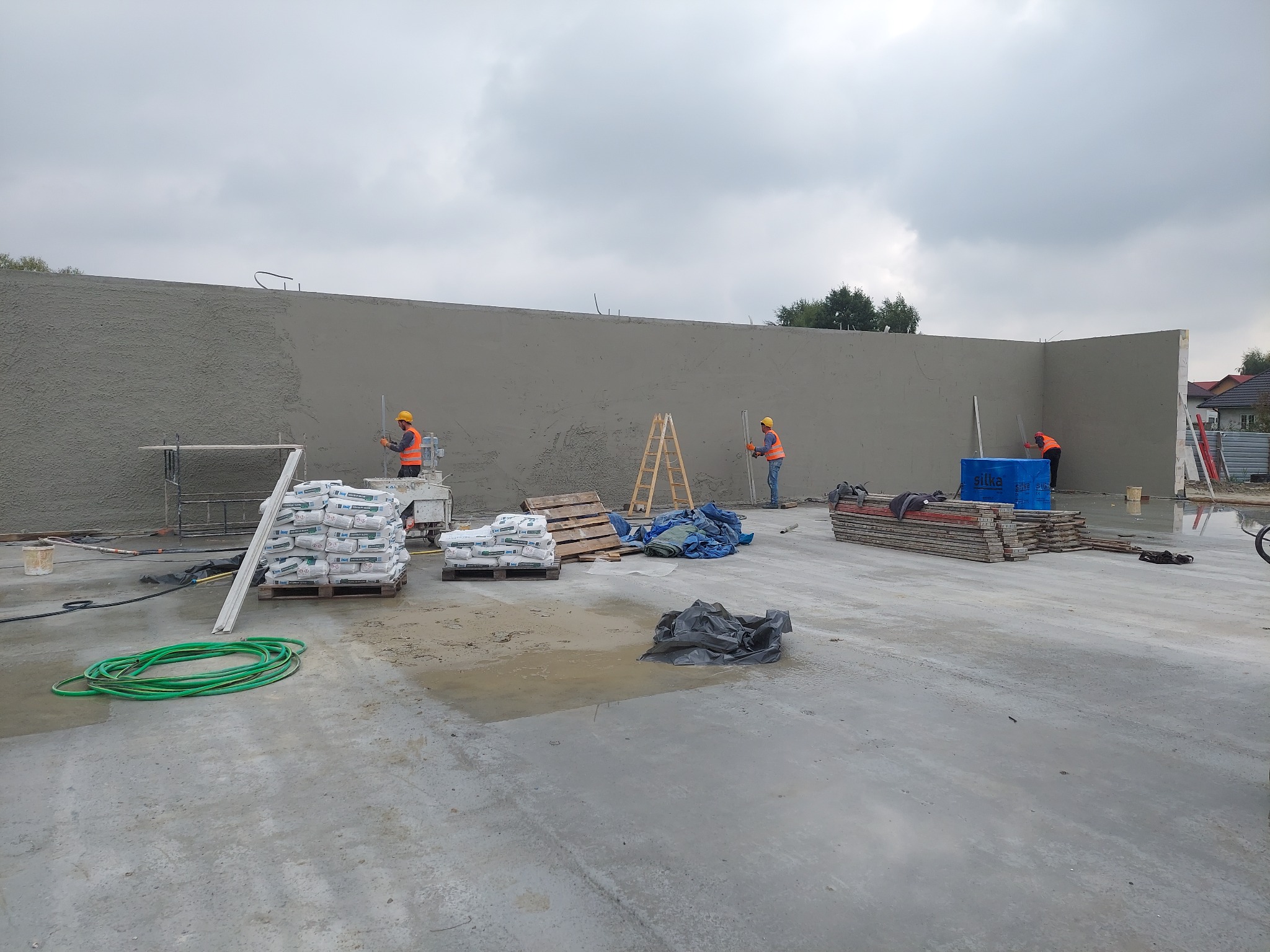 Trzech pracowników w pomarańczowych kamizelkach nakłada tynk na ścianę budynku. Na pierwszym planie palety z workami tynku, zielony wąż i kałuże na betonowej posadzce.