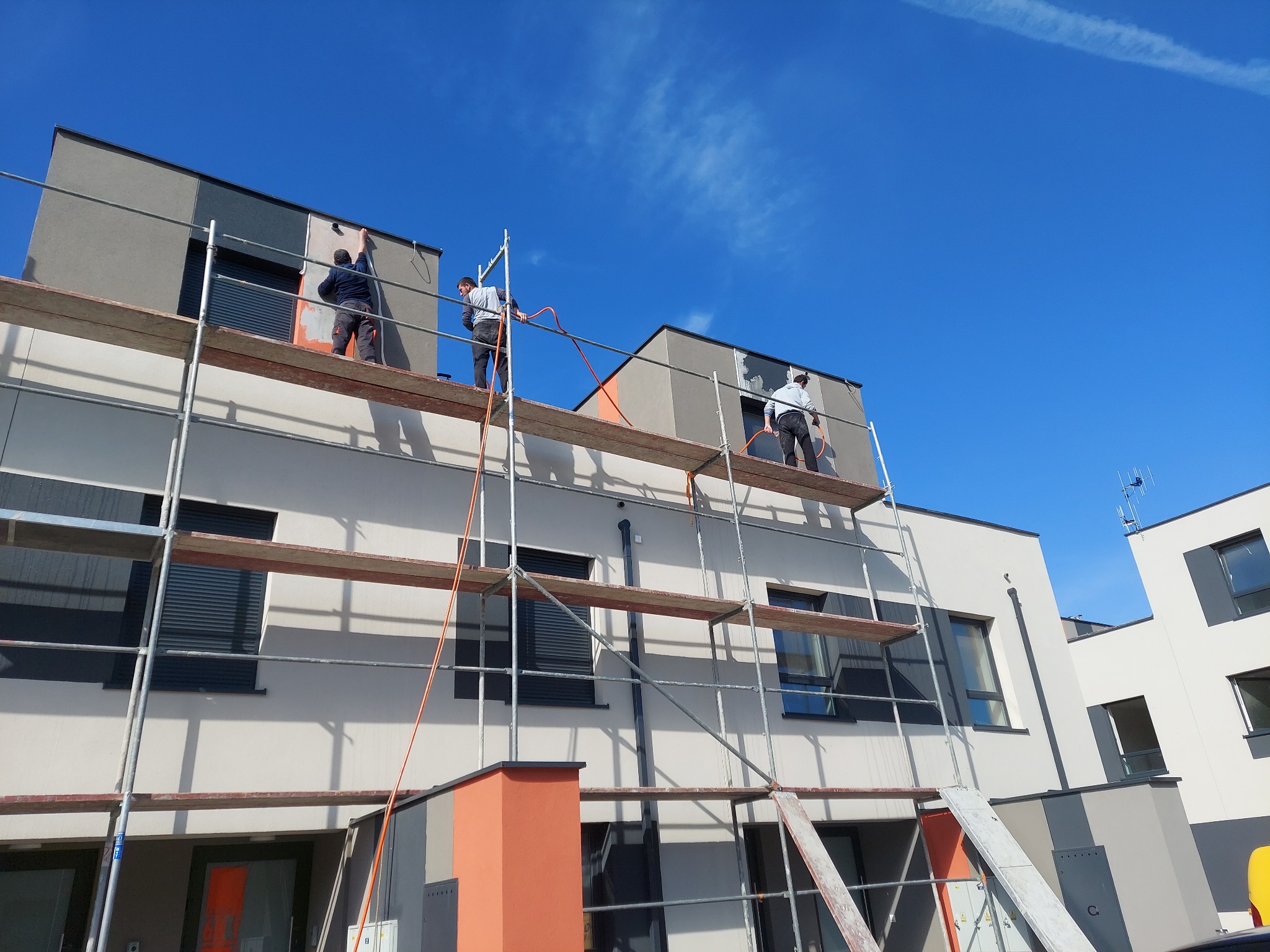 Ocieplanie i tynkowanie elewacji budynku wielorodzinnego z użyciem rusztowań, widoczni pracownicy podczas pracy, błękitne niebo w tle.