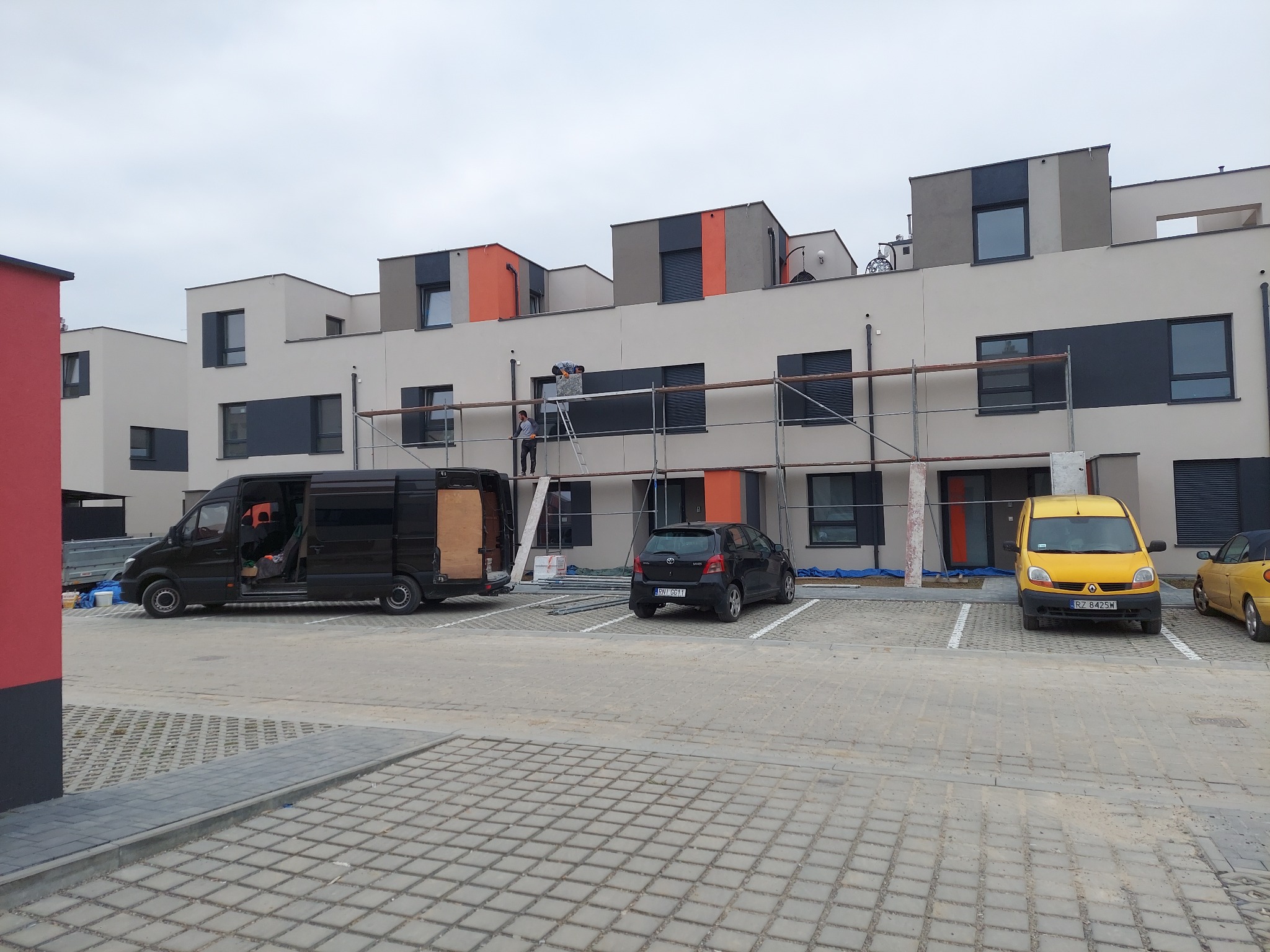Pracownicy na rusztowaniu przy elewacji budynku z elementami szarości i pomarańczy, zaparkowane samochody dostawcze i osobowe, kostka brukowa na parkingu.