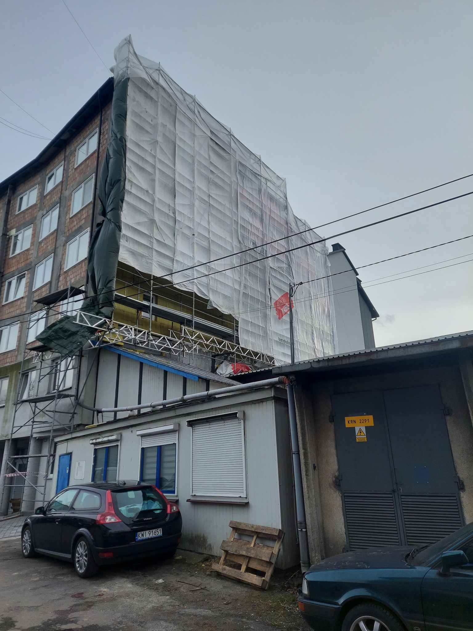 Wielopiętrowy budynek w trakcie prac elewacyjnych, osłonięty siatką ochronną, widoczne rusztowania, fragment ocieplenia oraz zaparkowane samochody w pobliżu.