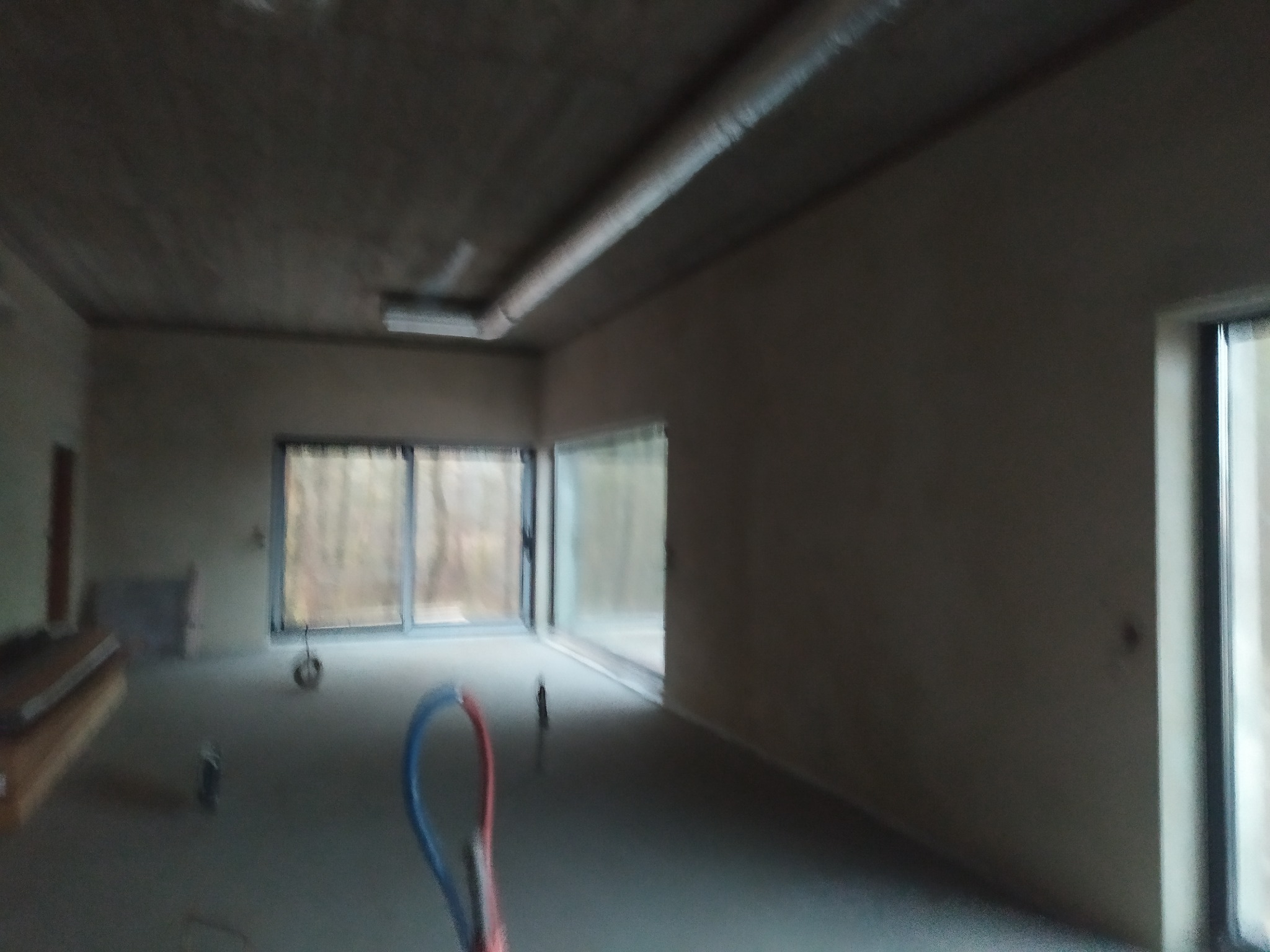 Wnętrze w trakcie prac wykończeniowych, widoczne ściany po tynkowaniu, rury instalacyjne na podłodze, duże okna z widokiem na las, surowy sufit z widoczną wentylacją.