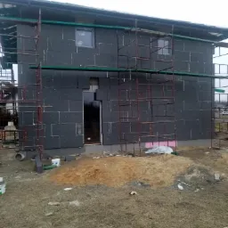 Dom w trakcie ocieplania elewacji, widoczne szare płyty izolacyjne, rusztowania i stos piasku.