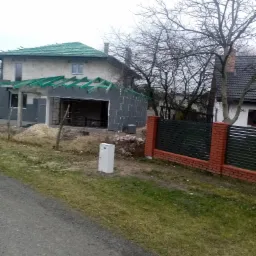 Surowy stan budowy domu jednorodzinnego z garażem, widoczne ściany z bloczków betonowych, zielona dachówka i niezakończona elewacja, na pierwszym planie skrzynka elektryczna.