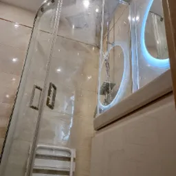 Zaokrąglona kabina prysznicowa z chromowanymi uchwytami i deszczownicą, widoczna składana ławeczka prysznicowa, umywalka z podświetlanym lustrem w łazience wykończonej beżowymi płytkami.