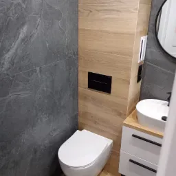 Nowoczesna łazienka z szarą, kamienną ścianą i drewnopodobnymi płytkami, wisząca toaleta z czarnym panelem, umywalka nablatowa na białej szafce z czarnymi uchwytami, okrągłe lustro.