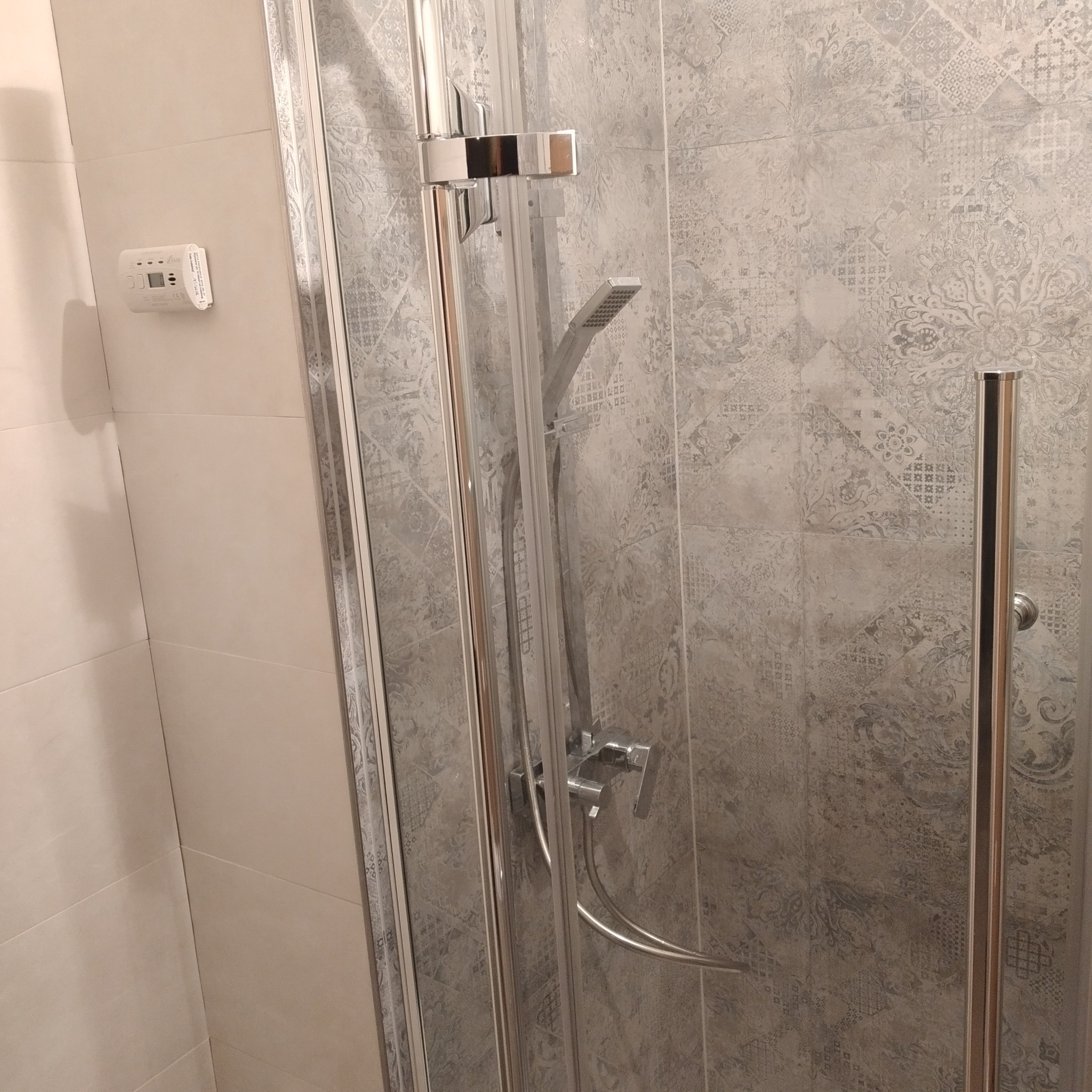 Kabina prysznicowa z chromowanymi elementami i dekoracyjnymi płytkami ściennymi w odcieniach szarości, zamontowany termostat na ścianie.