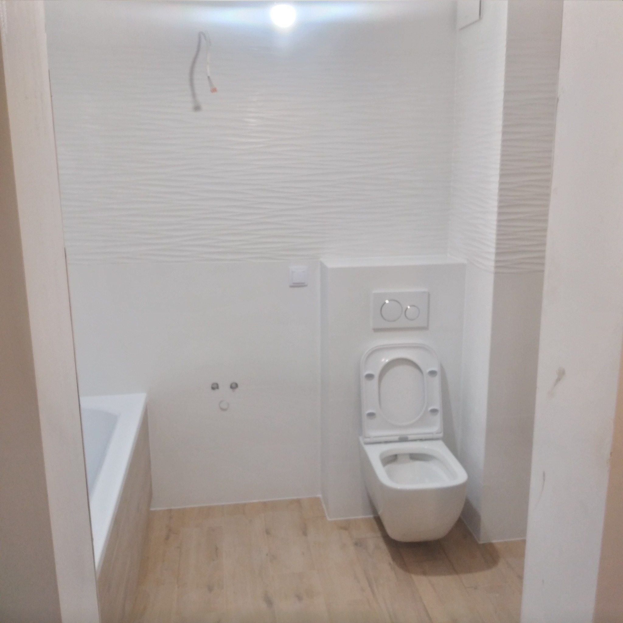 Nowoczesna, jasna łazienka po remoncie z podwieszaną toaletą, wanną i płytkami imitującymi fale na ścianach. Widoczne przygotowane wyprowadzenia instalacji.