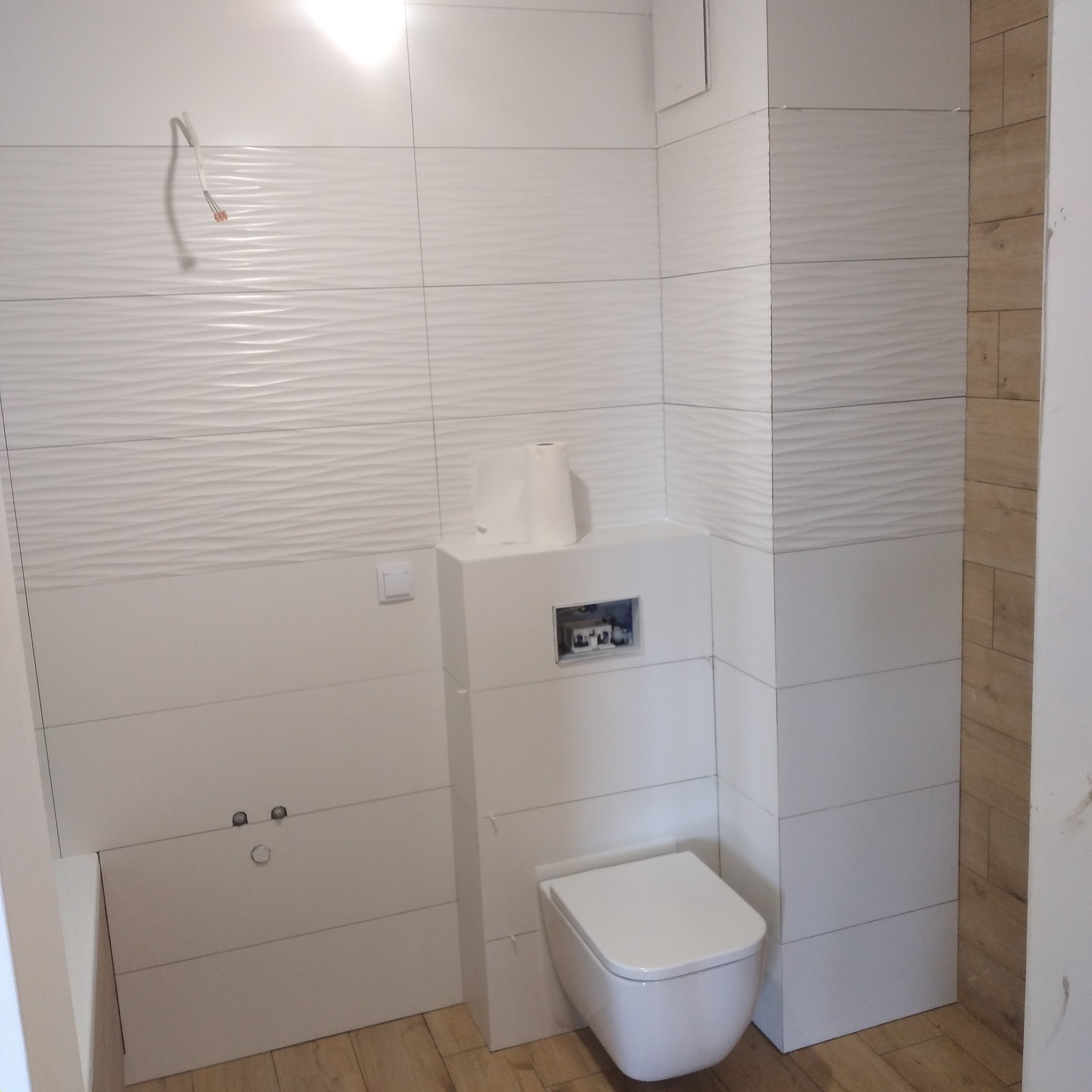 Nowoczesna łazienka w trakcie remontu, widoczne białe płytki z delikatnym wzorem, podwieszana toaleta, drewnopodobne płytki na podłodze i ścianie, wystające przewody elektryczne.