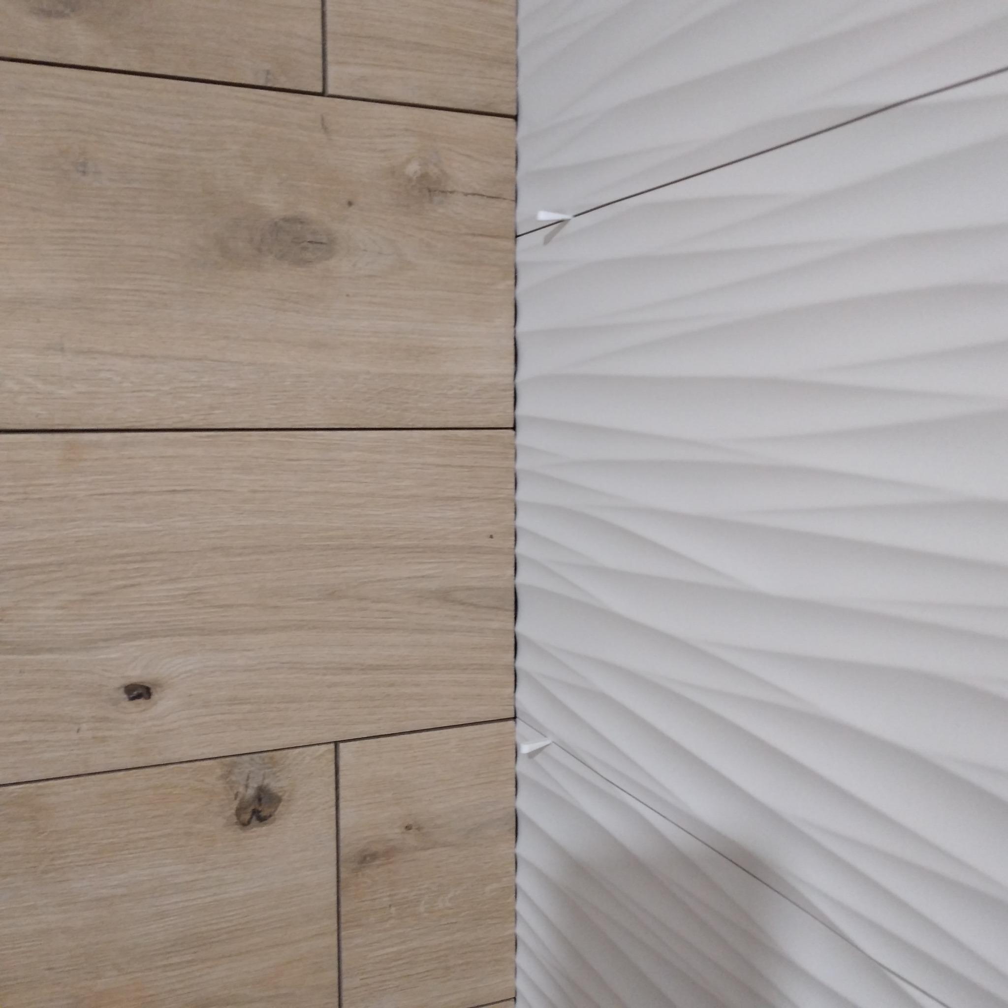 Połączenie płytek imitujących drewno i białych, trójwymiarowych płytek falistych na ścianie, widoczne krzyżyki dystansowe.